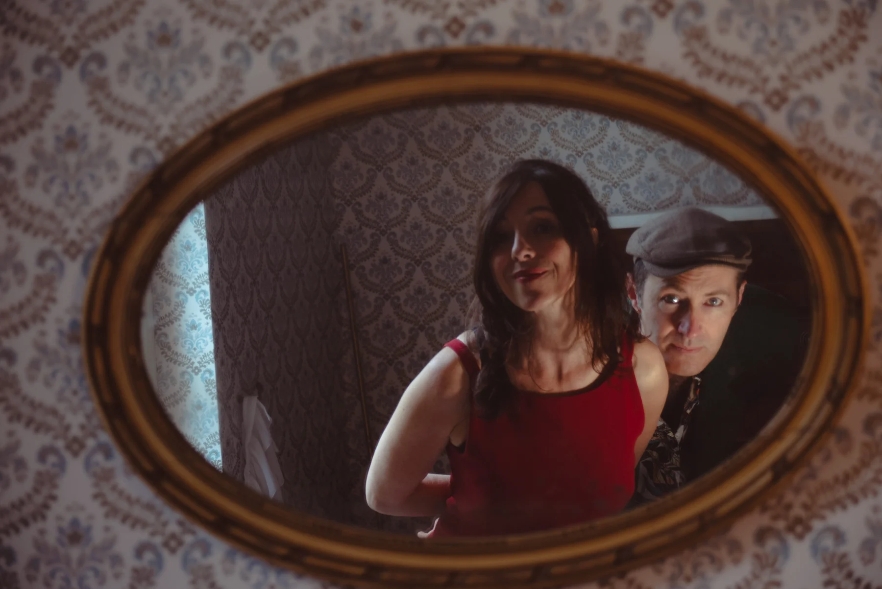 Sandra en robe rouge et Frédéric en casquette se reflètent dans un miroir ovale au cadre doré