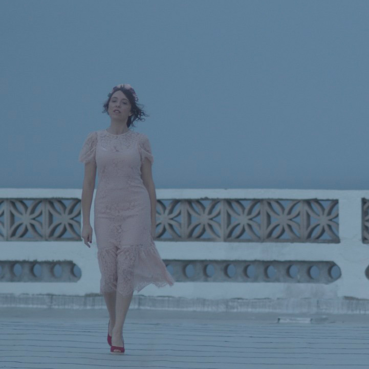 Sandra en robe claire et chaussures rouges sur une terrasse devant une balustrade et un ciel bleu